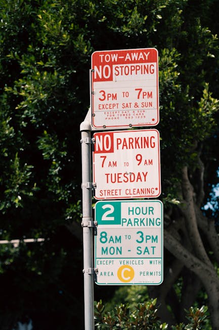The image shows a vertical metal pole with multiple parking and stopping regulation signs against a backdrop of lush green trees. The top sign is red and white, indicating a tow-away zone with no stop
