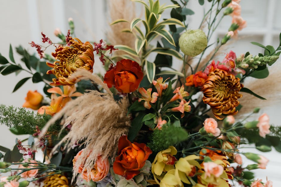 A vibrant floral arrangement featuring orange, yellow, and pink roses, complemented by large protea flowers with reddish-brown petals, delicate pink blossoms, and feathery pampas grass. The bouquet is