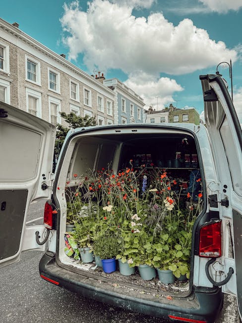 The back of a white van parked on a city street, filled with potted plants and flowers including red poppies, white daisies, and green foliage, ready for flower delivery or floral arrangement. The veh