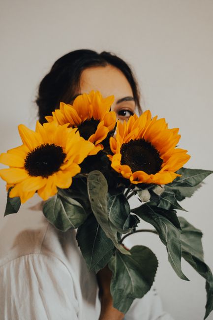 A person holding a vibrant bouquet of large, bright yellow sunflowers with dark centers and lush green leaves, partially obscuring their face. The bouquet appears freshly arranged, featuring sturdy st