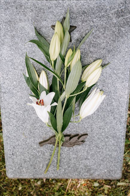 A delicate floral tribute featuring a small arrangement of white lilies and white lilies with closed buds, arranged on a grey stone memorial. The lilies have large, trumpet-shaped petals with smooth, 