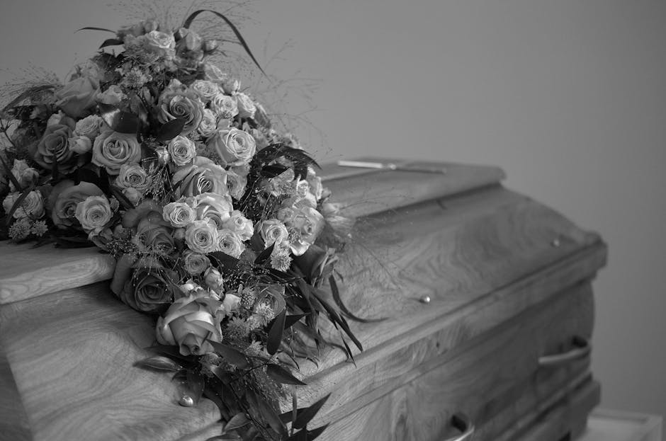 A large, overflowing funeral bouquet arranged with a variety of fresh roses in different sizes and shades, predominantly light-colored and some darker roses, set on a wooden coffin. The bouquet includ