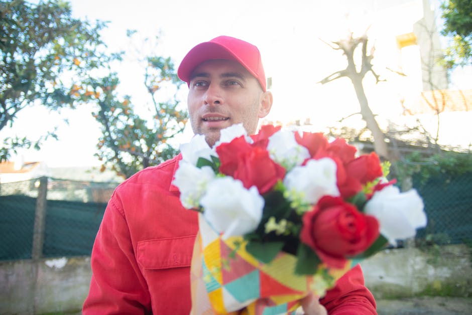 A man dressed in a red jacket and matching red cap holds out a colorful bouquet of fresh flowers towards the camera. The bouquet contains red and white roses arranged with green foliage, wrapped in de