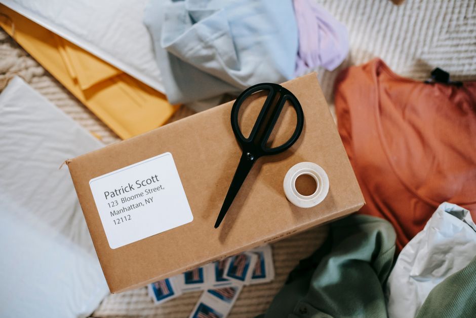 A cardboard parcel with a white label displaying the name 'Patrick Scott' and an address in Manhattan, NY, lies on a beige surface surrounded by various wrapping materials such as tissue paper and env