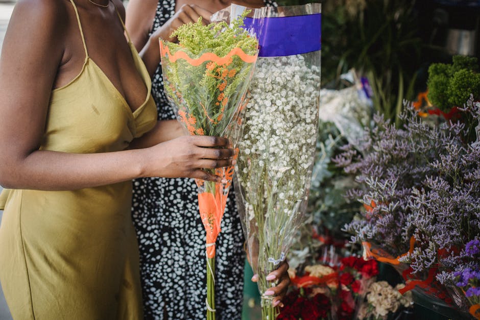 A woman in a yellow spaghetti strap dress holding a large bouquet of green and white flowers wrapped in clear plastic with orange decorative paper at the base. The bouquet contains small green cluster